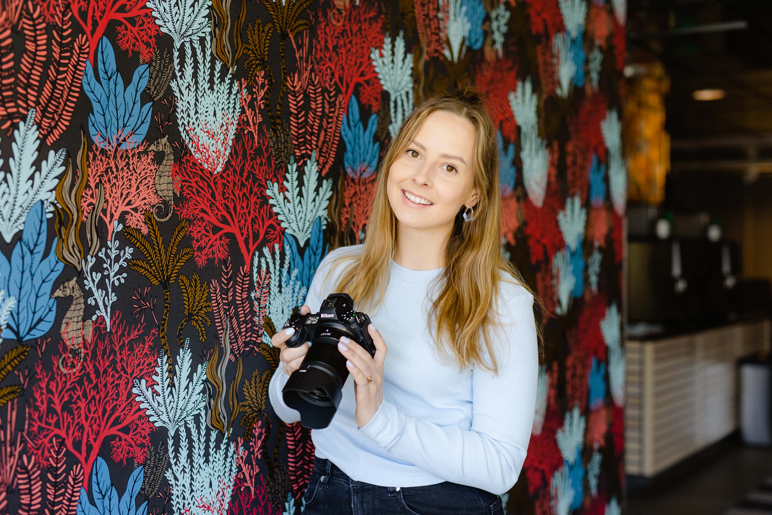 Portrett av fotograf Anne Katja Gaup som smiler til kamera mens hun står mot en rød vegg.