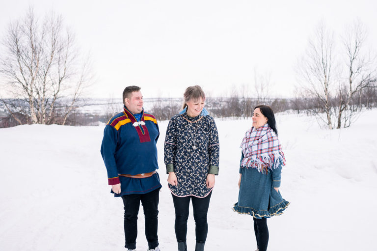 Tre samiske ansatte står ute i snøen og smiler til hverandre.